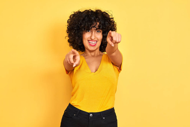 Young arab woman with curly hair wearing t-shirt standing over isolated yellow background pointing to you and the camera with fingers, smiling positive and cheerful - Photo, Image