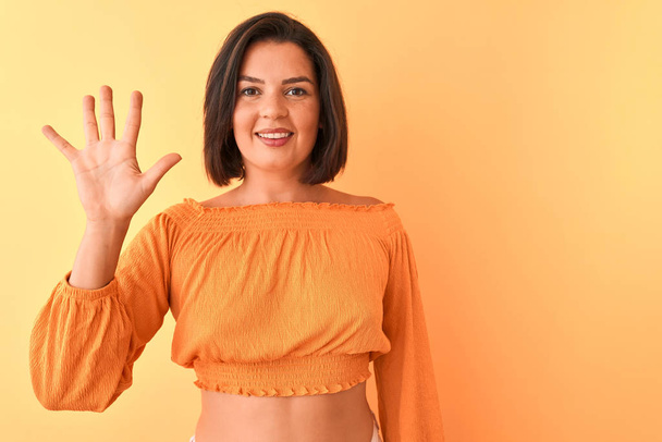 Νεαρή όμορφη γυναίκα φορώντας casual t-shirt στέκεται πάνω από απομονωμένο πορτοκαλί φόντο που δείχνει και δείχνει προς τα πάνω με τα δάχτυλα νούμερο πέντε, ενώ χαμογελά αυτοπεποίθηση και χαρούμενος. - Φωτογραφία, εικόνα