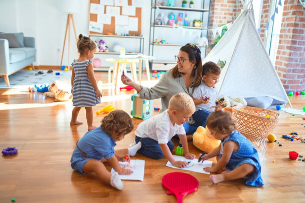 Hermosa maestra y grupo de niños pequeños sentados en el piso dibujando con papel y lápiz alrededor de muchos juguetes en el jardín de infantes
 - Foto, imagen