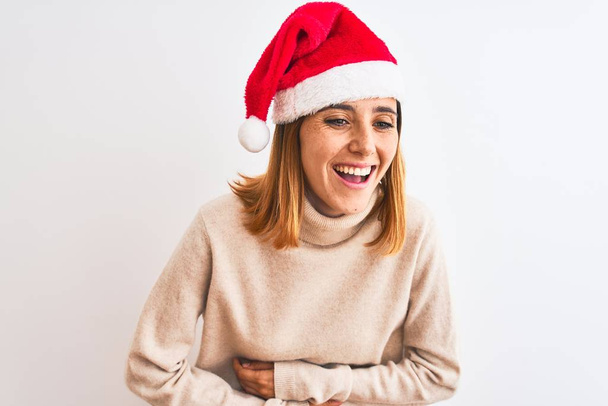 Hermosa pelirroja con sombrero de navidad sobre fondo aislado sonriendo y riendo en voz alta porque divertida broma loca con las manos en el cuerpo
. - Foto, Imagen