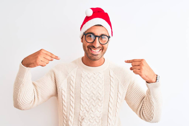 Young handsome man wearing christmas hat and glasses over isolated background looking confident with smile on face, pointing oneself with fingers proud and happy. - Foto, Imagen
