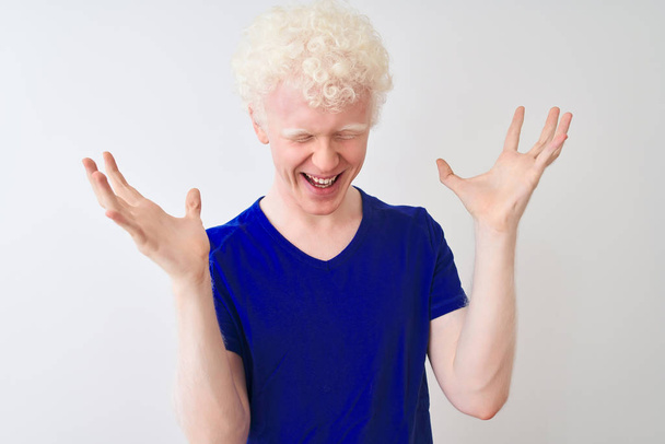 Jonge albino blonde man draagt blauwe casual t-shirt staan over geïsoleerde witte achtergrond vieren gek en gek voor succes met armen omhoog en gesloten ogen schreeuwen opgewonden. Winnaars concept - Foto, afbeelding