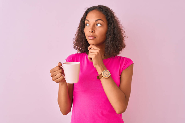 Young brazilian woman drinking cup of coffee standing over isolated pink background serious face thinking about question, very confused idea - Foto, imagen