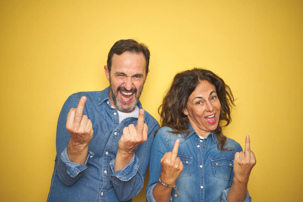 Beautiful middle age couple together standing over isolated yellow background Showing middle finger doing fuck you bad expression, provocation and rude attitude. Screaming excited - Photo, Image