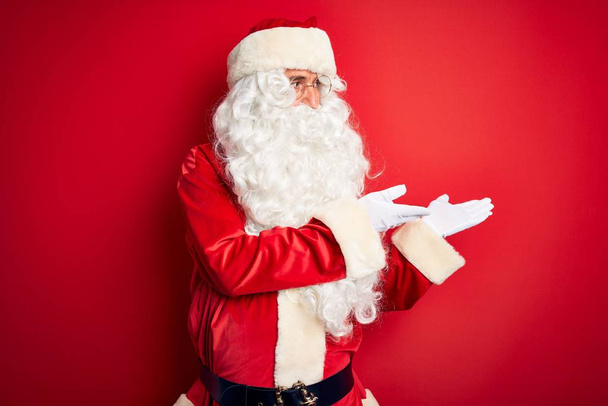 gutaussehender Mann mittleren Alters im Weihnachtsmannkostüm, der vor isoliertem roten Hintergrund steht und zur Seite zeigt, mit den Händen geöffnete Handflächen, die Kopierraum zeigen, und Werbung präsentiert, die aufgeregt glücklich lächelt - Foto, Bild