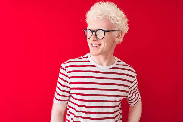 Jonge albino blonde man met gestreept t-shirt en een bril over geïsoleerde rode achtergrond die van links naar rechts kijkt met een glimlach op het gezicht, natuurlijke expressie. Lachend vol vertrouwen. - Foto, afbeelding