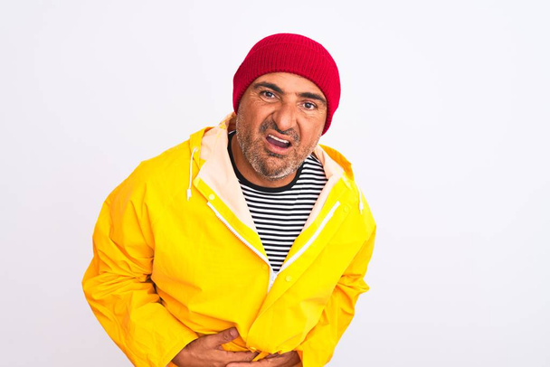 Middle age man wearing rain coat and woolen hat standing over isolated white background with hand on stomach because indigestion, painful illness feeling unwell. Ache concept. - Photo, Image