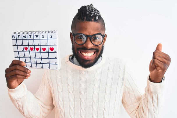 Afrikanisch-amerikanischer Mann mit Zöpfen, die Zeitrahmen-Kalender vor isoliertem weißen Hintergrund halten, schreien stolz und feiern Sieg und Erfolg sehr aufgeregt, jubelnde Emotion - Foto, Bild
