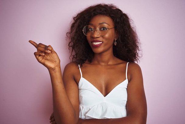 Jonge Afro-Amerikaanse vrouw met een bril over een geïsoleerde roze achtergrond met een grote glimlach op haar gezicht, wijzend met hand en vinger naar de zijkant kijkend naar de camera. - Foto, afbeelding