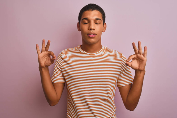Jeune homme arabe beau portant t-shirt rayé debout sur fond rose isolé se détendre et sourire les yeux fermés en faisant geste de méditation avec les doigts. Concept de yoga
. - Photo, image