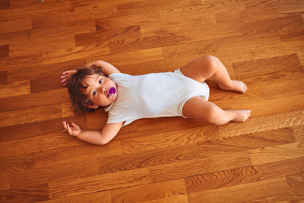 Όμορφο μικρό κοριτσάκι φορώντας λευκό κοστούμι του σώματος ξαπλωμένο στο πάτωμα χρησιμοποιώντας πιπίλα - Φωτογραφία, εικόνα