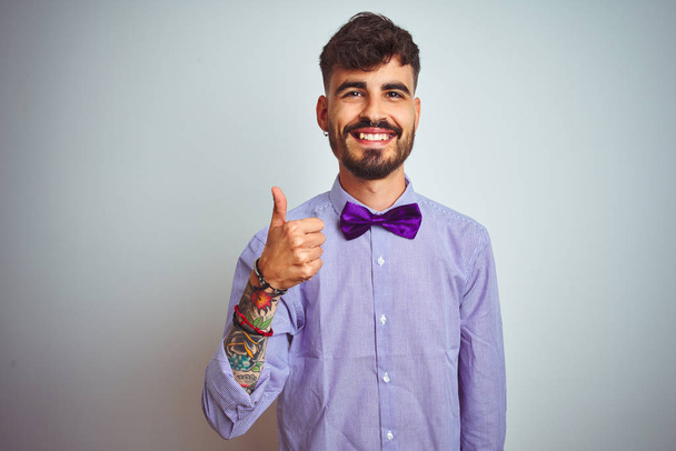 Jeune homme avec tatouage portant chemise violette et noeud papillon sur fond blanc isolé faisant des pouces heureux geste avec la main. Approuver l'expression en regardant la caméra montrant le succès
. - Photo, image