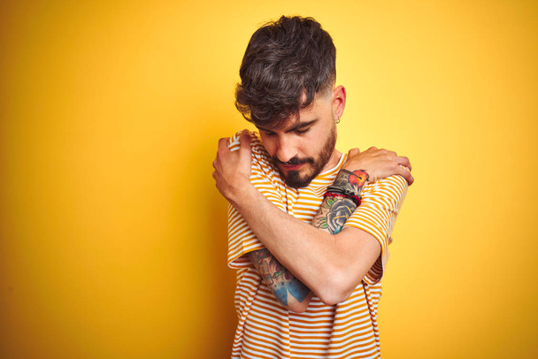 Mladý muž s tetováním v pruhovaném tričku, stojící nad osamělým žlutým pozadím, objímající se šťastný a pozitivní, usmívající se sebevědomě. Sebeláska a sebepéče - Fotografie, Obrázek