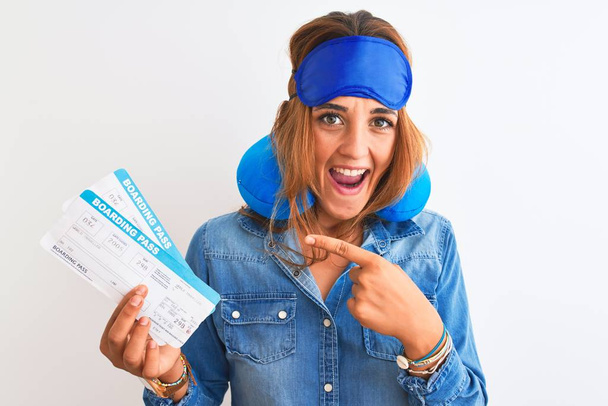 Young redhead tourist woman wearing sleeping mask and neck pillow showing boarding pass very happy pointing with hand and finger - Photo, Image