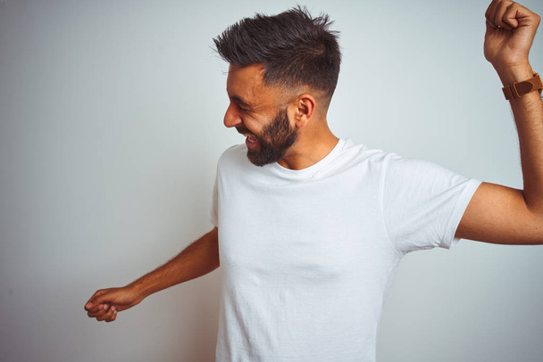 Young indian man wearing t-shirt standing over isolated white background Dancing happy and cheerful, smiling moving casual and confident listening to music - Foto, immagini