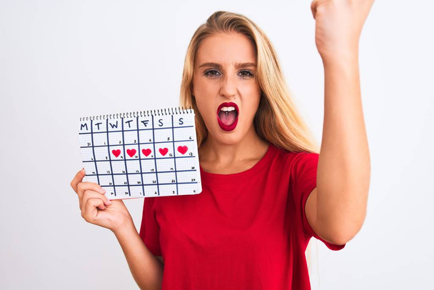 Young beautiful woman holding period calendar standing over isolated white background annoyed and frustrated shouting with anger, crazy and yelling with raised hand, anger concept - 写真・画像