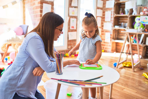 Καυκάσιο κορίτσι παίζει και μαθαίνει στο σχολείο με μια δασκάλα. Μητέρα και κόρη στο playroom γύρω από παιχνίδια ζωγραφισμένα σε μαγνητικό μαυροπίνακα - Φωτογραφία, εικόνα