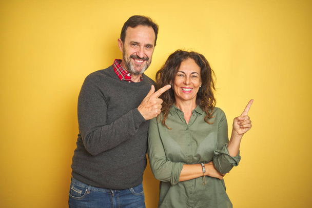 Όμορφη μεσήλικη ζευγάρι πάνω από απομονωμένο κίτρινο φόντο με ένα μεγάλο χαμόγελο στο πρόσωπο, δείχνοντας με το χέρι και το δάχτυλο στην πλευρά κοιτάζοντας την κάμερα. - Φωτογραφία, εικόνα