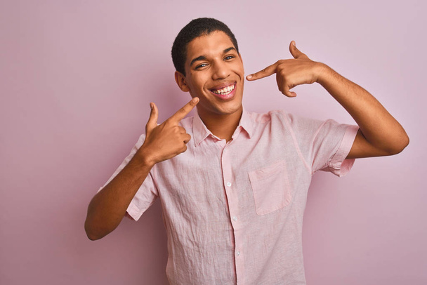 Nuori komea arabi mies päällään rento paita seisoo yli eristetty vaaleanpunainen tausta hymyilevä iloinen osoittaa ja sormilla hampaat ja suu. Hammasterveyden käsite
. - Valokuva, kuva