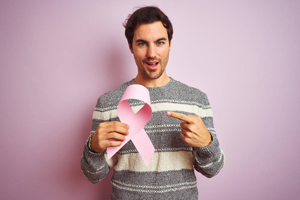 Młody przystojny mężczyzna trzyma wstążkę raka stojąc nad odizolowanym różowym tle bardzo szczęśliwy wskazując ręką i palcem - Zdjęcie, obraz