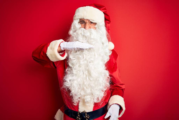 Pohledný muž středního věku v kostýmu Santa stojí nad izolovaným červeným pozadím, podřezává hrdlo rukou jako nůž, hrozí agresivitou zuřivým násilím - Fotografie, Obrázek