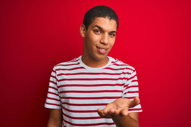 Jonge knappe arabische man draagt gestreept t-shirt over geïsoleerde rode achtergrond glimlachend vrolijk aanbieden palm hand geven hulp en acceptatie. - Foto, afbeelding