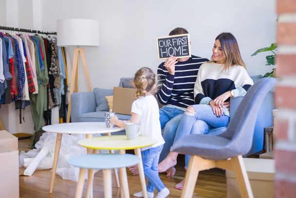 Hermosa familia con niño sentado en el sofá sosteniendo pizarra en el nuevo hogar alrededor de cajas de cartón
 - Foto, Imagen