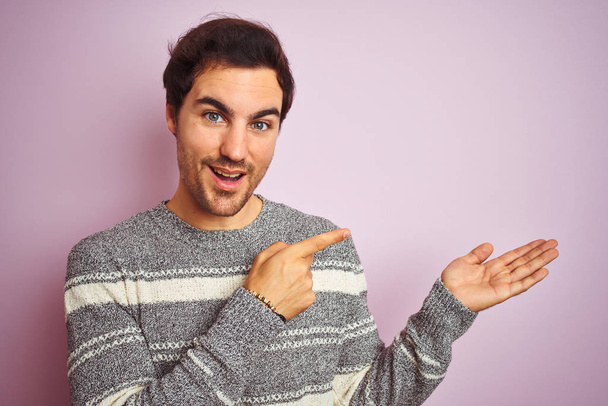 Joven hombre guapo con suéter a rayas casual de pie sobre fondo rosa aislado muy feliz señalando con la mano y el dedo
 - Foto, imagen