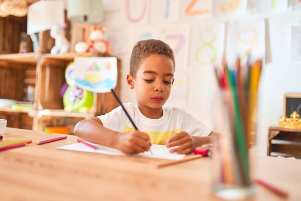 Beau bambin afro-américain assis à dessiner en utilisant du papier et des crayons sur le bureau à la maternelle
 - Photo, image