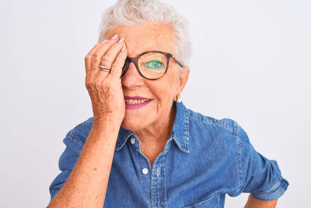 Senior harmaa tukka nainen yllään denim paita ja lasit yli eristetty valkoinen tausta peittää toinen silmä käsin, luottavainen hymy kasvoilla ja yllätys tunteita
. - Valokuva, kuva