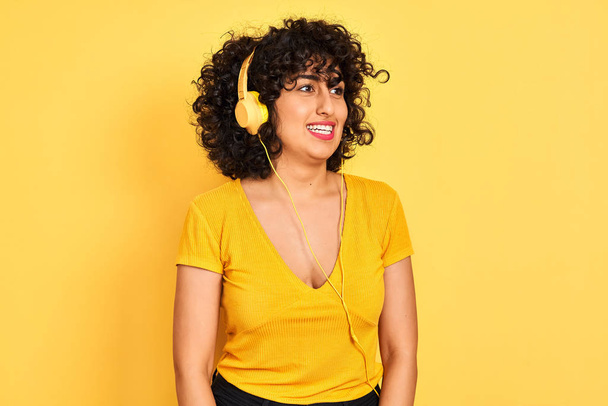Arabka s kudrnatými vlasy, která poslouchá hudbu pomocí sluchátek na izolovaném žlutém pozadí, dívá se stranou s úsměvem na tváři, přirozený výraz. Smějící se sebevědomí. - Fotografie, Obrázek