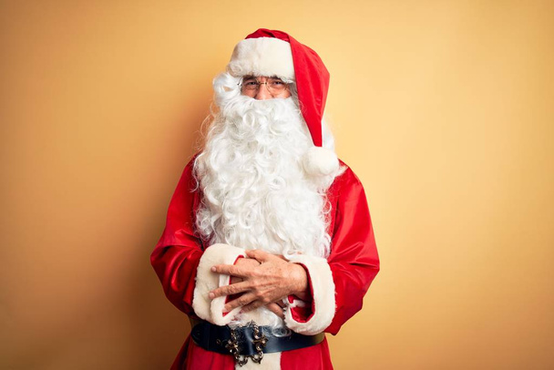 Orta yaşlı yakışıklı bir adam, Noel Baba kostümü giymiş, izole edilmiş sarı arka plan üzerinde durmuş kollarını kavuşturup kameraya bakarken gülümsüyor. Pozitif kişi. - Fotoğraf, Görsel