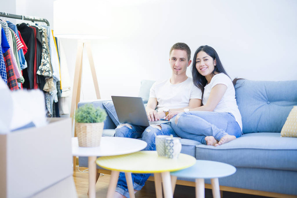 Young beautiful couple sitting on the sofa using laptop at new home around cardboard boxes - Foto, imagen