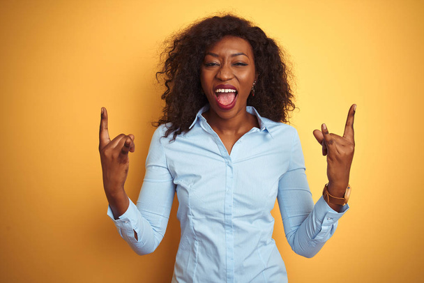 Femme d'affaires afro-américaine portant une chemise élégante sur fond jaune isolé criant avec une expression folle faisant symbole rock avec les mains en l'air. Une star de la musique. Concept lourd
. - Photo, image