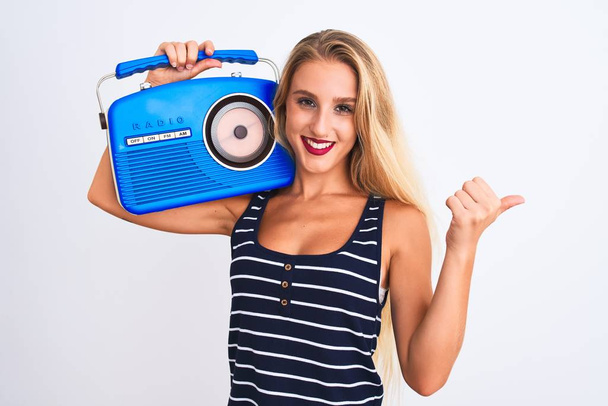 Young beautiful woman holding vintage radio standing over isolated white background pointing and showing with thumb up to the side with happy face smiling - Zdjęcie, obraz