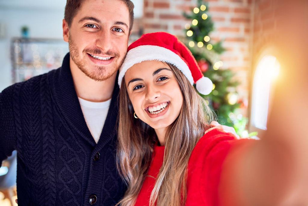 Young beautiful couple smiling happy and confident. Standing make selfie by camera around christmas tree at home - Фото, изображение