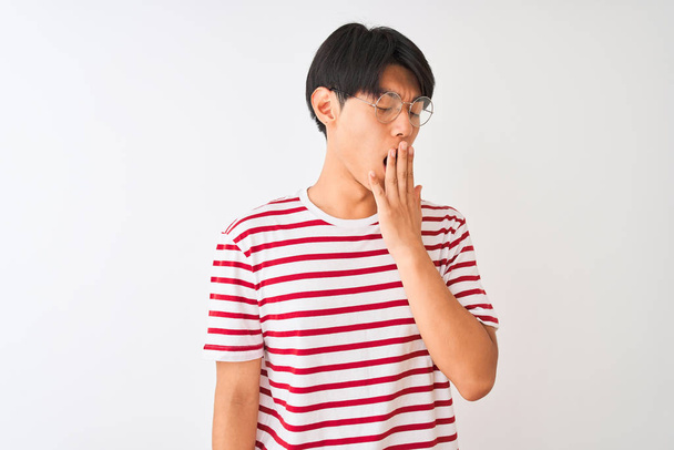 Joven hombre chino con gafas y camiseta a rayas de pie sobre fondo blanco aislado aburrido bostezo cansado cubriendo la boca con la mano. Inquietud y somnolencia
. - Foto, imagen