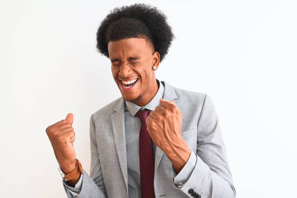 Mladý africký americký podnikatel v obleku, stojící nad osamělým bílým pozadím, velmi šťastný a vzrušený, dělá vítězný gesto se zdviženýma rukama, usmívá se a křičí po úspěchu. Koncept oslavy. - Fotografie, Obrázek