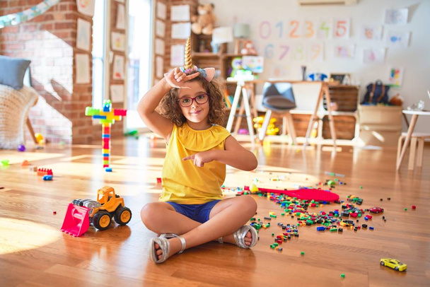 Beautiful toddler wearing glasses and unicorn diadem sitting on the floor at kindergarten smiling making frame with hands and fingers with happy face. Creativity and photography concept. - Photo, Image