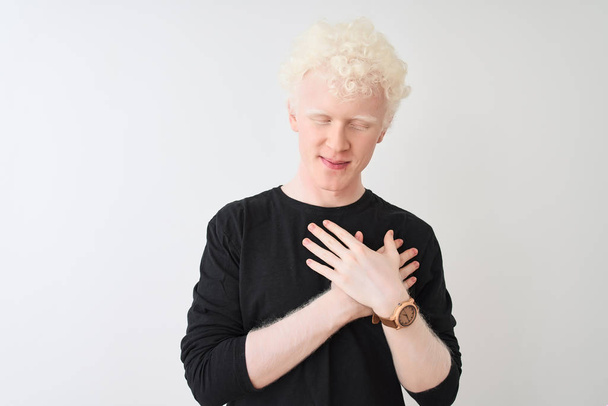 Mladý blonďatý albín v černém tričku, stojící nad osamělým bílým pozadím, usmívající se s rukama na prsou se zavřenýma očima a vděčným gestem na tváři. Zdravotní koncepce. - Fotografie, Obrázek