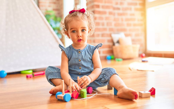 Piękne białe niemowlę bawiące się zabawkami w kolorowym pokoju zabaw. Szczęśliwy i zabawny z drewnianymi elementami pociągu w przedszkolu. - Zdjęcie, obraz