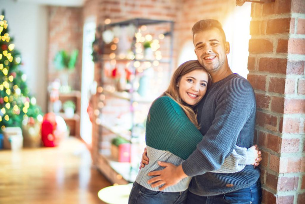 Young beautiful couple smiling happy and confident. Standing and hugging around christmas tree at home - Foto, afbeelding