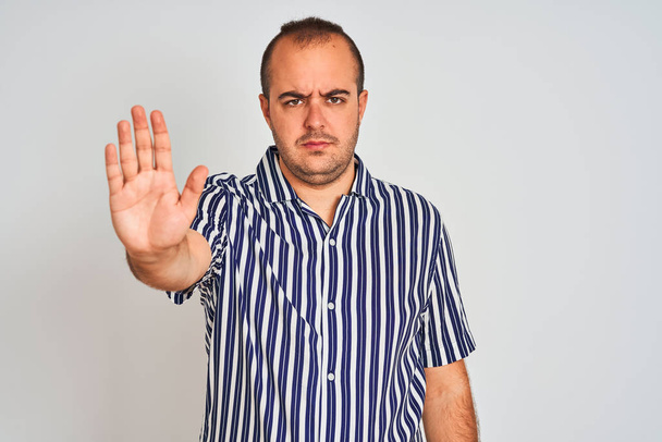 Νεαρός άνδρας φορώντας μπλε ριγέ πουκάμισο στέκεται πάνω από απομονωμένο λευκό φόντο κάνει σταματήσει να τραγουδά με την παλάμη του χεριού. Προειδοποιητική έκφραση με αρνητική και σοβαρή χειρονομία στο πρόσωπο. - Φωτογραφία, εικόνα