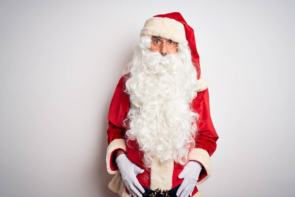 Middle age handsome man wearing Santa costume standing over isolated white background In shock face, looking skeptical and sarcastic, surprised with open mouth - Foto, imagen