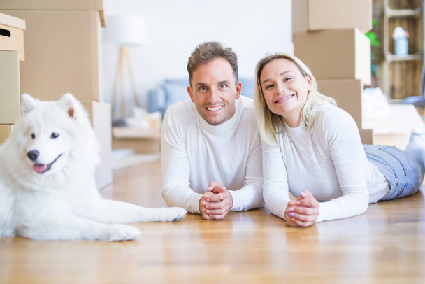 Young beautiful couple with dog lying down on the floor at new home around cardboard boxes - Photo, Image
