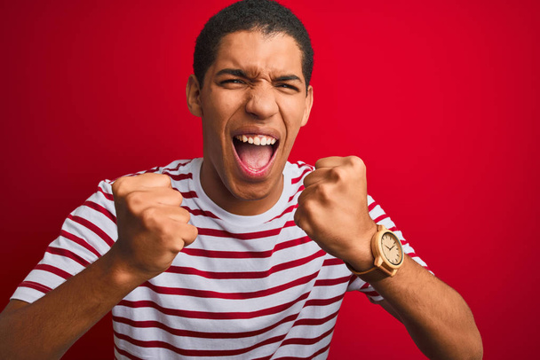 Joven hombre árabe guapo con camiseta a rayas sobre fondo rojo aislado enojado y loco levantando puños frustrados y furiosos mientras gritaba con ira. Rabia y concepto agresivo
. - Foto, imagen