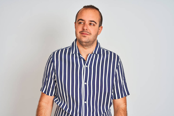 Νεαρός άνδρας φορώντας μπλε ριγέ πουκάμισο στέκεται πάνω από απομονωμένο λευκό φόντο χαμογελώντας κοιτάζοντας στο πλάι και κοιτάζοντας μακριά σκέψης. - Φωτογραφία, εικόνα
