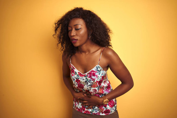 消化不良、痛みを伴う病気が具合が悪いので、アフリカ系アメリカ人女性は胃の上に手で隔離された黄色の背景に花の夏のTシャツを着ています。アチェの概念. - 写真・画像