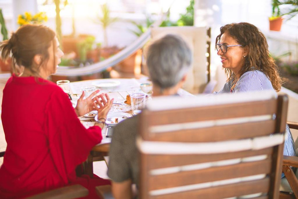 Встреча женщин среднего возраста, обедающих и пьющих кофе. Зрелые друзья улыбаются счастливо, используя смартфон дома в солнечный день
 - Фото, изображение