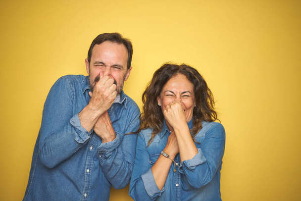 Beautiful middle age couple together standing over isolated yellow background smelling something stinky and disgusting, intolerable smell, holding breath with fingers on nose. Bad smells concept. - Photo, Image